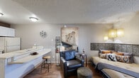 This thoughtfully arranged studio offers a seamless blend of living and functional spaces across a single floor. The combined kitchen and living area serves as the heart of the unit, where a well-equipped kitchen features white cabinetry with ample storage, light countertops, a full-size refrigerator, an electric range, and a double sink — all set against a warm, contrasting accent wall. A peninsula counter extends into the living space, offering additional workspace and casual seating with an industrial-style stool.

The living area is warm and inviting, with rich wood-look flooring that flows throughout the entire unit. A large window fitted with vertical blinds allows natural light to fill the room, brightening the textured ceiling above. The space accommodates a sleeping area, a cozy leather lounge chair, and a compact desk nook, making it highly functional for both rest and work.

Pendant lighting adds a stylish touch over the living zone, complementing the urban-inspired décor. The bathroom is clean and straightforward, featuring wood-look flooring, a vanity with sink, and a shower enclosed by a curtain. A dedicated closet provides practical storage. Overall, the studio presents a cozy, urban-style living environment with efficient use of every square foot.