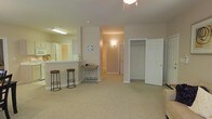 Located at 1530 Wimbledon Dr in Greenville, NC, this single-floor residence offers a well-designed, open layout that maximizes livability and flow throughout. The main living area is finished with plush wall-to-wall carpeting in a warm neutral tone, creating a comfortable and inviting atmosphere. A peninsula breakfast bar with seating separates the kitchen from the living space, making it ideal for casual dining or entertaining. The kitchen features white cabinetry and is reflected in a mirror accent, adding depth and brightness to the area. Neutral-toned walls throughout lend a clean, cohesive aesthetic that complements a variety of décor styles. A hallway leads to additional rooms, offering a sense of privacy and separation between living and sleeping areas. A spacious closet with double-door access provides ample storage, thoughtfully integrated into the living space. Overhead lighting fixtures illuminate the home efficiently, while the overall layout ensures easy navigation between spaces. The single-floor design makes this home highly accessible, with all living areas conveniently located on one level. This residence combines functional design with comfortable finishes, making it a practical and welcoming home in the Greenville, NC area.