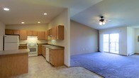 This single-floor space offers a practical and comfortable open-concept layout that seamlessly connects the kitchen and living area. The kitchen is equipped with a white electric range, dishwasher, and a double-basin sink, all complemented by warm medium-toned wood cabinetry that provides generous storage space. Dark countertops offer a subtle contrast to the light-colored cabinetry, while recessed lighting illuminates the workspace effectively. The flooring transitions from easy-to-clean vinyl tile in the kitchen to plush carpeting in the living area, creating a cozy and defined separation between the two spaces. The living area is highlighted by a ceiling fan with a light fixture, ensuring year-round comfort. A sliding glass door bathes the living space in natural light and provides direct access to a private balcony, extending the usable living space outdoors. Neutral wall tones throughout the unit create a clean, versatile backdrop suitable for a variety of interior styles. The open flow between the kitchen and living area makes the space feel inviting and functional, ideal for both everyday living and casual entertaining.