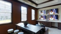 This single-floor space is designed with a sophisticated and inviting aesthetic, blending warm, earthy tones with refined architectural details. The walls are finished in a deep, rich brown hue, complemented by dark wood crown molding and trim that frame the room with an elegant contrast against the cream-colored tray ceiling above. Recessed lighting adds a polished touch to the overhead design.

Natural light pours in through multiple tall, double-hung windows, brightening the space and offering views of the surrounding exterior. A large, rustic dark wood communal table anchors the room, accompanied by round stools, making it well-suited for gatherings or collaborative use. A secondary round accent table nearby adds to the layered seating arrangement.

A key feature of the space is the built-in dark wood cabinetry along one wall, which serves as a functional console base for two wall-mounted flat-screen televisions arranged in a stacked configuration — ideal for viewing events or presentations simultaneously. Decorative accents, including a centerpiece floral arrangement and styled trays, add warmth and character throughout. Overall, the space strikes a balance between functional utility and upscale design.