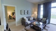 This single-floor residence offers a cohesive and comfortable living environment with a clean, modern aesthetic. The living area serves as the heart of the home, finished in soft blue-gray tones that create a calm and inviting atmosphere. Plush carpeted flooring runs throughout, adding warmth underfoot. The space is furnished with a gray sofa accented by decorative throw pillows, a glass-topped coffee table, and a coordinating armchair, all arranged to encourage relaxed conversation. A trio of framed artwork adorns the main wall, complementing the understated color palette, while a sleek floor lamp provides warm ambient lighting. Natural light flows generously through a large window dressed with elegant gray grapevine curtains, offering views of the lush greenery outside. An open passageway connects the living area seamlessly to the bedroom, which features a warm yellow accent wall, a richly upholstered headboard, and cozy bedding, creating a distinct yet harmonious transition between spaces. The overall design balances functionality with comfort, making thoughtful use of the available floor plan to deliver a bright, well-appointed, and livable environment.