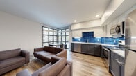 This single-floor unit in Berkeley, California offers a thoughtfully designed layout that balances communal living with private space. The open-concept living and kitchen area serves as the heart of the home, anchored by a striking kitchen featuring two-tone cabinetry—white uppers paired with charcoal lowers—and a vivid cobalt blue subway tile backsplash that adds a bold visual accent. Stainless steel appliances, including a range, microwave, dishwasher, and refrigerator, complement the white quartz countertops. Light wood-look flooring runs throughout the main living areas, enhancing the bright, airy atmosphere.

A floor-to-ceiling corner window arrangement floods the dining nook with natural light and outdoor views, seamlessly connecting the interior to its surroundings. The secondary living area provides additional lounge space with hardwood-style flooring and a television setup.

The bedrooms feature plush carpeting, window blinds, and functional study desks, accommodating multiple occupants comfortably. The clean, minimalist bathroom includes a soaking tub, shower, and modern white vanity with wood flooring. A separate vanity area sits adjacent to a conveniently located in-unit stacked washer and dryer, adding everyday practicality to this well-appointed space.