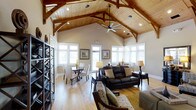 This single-story space makes a strong architectural statement through its soaring vaulted ceiling, clad in warm tongue-and-groove pine planking and supported by richly finished exposed timber trusses. The structural beams add a distinctive craftsman character, while recessed lighting and a large ceiling fan ensure both functionality and comfort throughout the space.

The open layout seamlessly integrates a living area and dining zone. The living area is anchored by a dark leather sofa and accent seating arranged over a patterned area rug, creating an inviting focal point. A dark wood bookshelf unit lines one wall, offering both storage and display space. The dining area features a dark wood table surrounded by upholstered chairs, positioned to take advantage of the natural light.

Abundant windows along multiple walls flood the interior with natural light and provide views to the outdoors, enhancing the sense of openness. Light hardwood flooring runs throughout, complementing the warm tones of the ceiling and timber work. The overall ambiance strikes a balance between rustic architectural detail and comfortable, well-appointed furnishings.