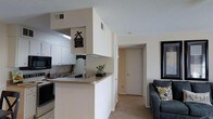 This single-floor residence offers a well-organized and efficient layout designed to maximize comfort and functionality. The kitchen is outfitted with white cabinetry that extends to the ceiling, providing generous storage space. A built-in overhead cabinet structure frames the kitchen area, creating a defined boundary while maintaining an open feel. The kitchen features a stainless steel sink, an electric range, and a refrigerator, all neatly arranged along the wall. A peninsula breakfast bar with a light countertop extends outward, offering a convenient casual dining or prep space and serving as a natural transition into the living area. The flooring transitions from tile in the kitchen to plush carpeting in the living and hallway areas, adding warmth underfoot. The living room is visible from the kitchen, furnished with a comfortable sofa and accented with decorative throw pillows, creating a cozy, inviting atmosphere. Neutral wall tones throughout the space lend a clean, airy quality. A hallway leads to a door at the rear, suggesting additional rooms or a private entrance. Textured ceilings and ceiling-mounted HVAC vents are consistent throughout, reflecting a practical residential design.