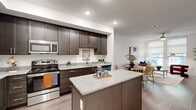 This thoughtfully designed 1-bedroom, 1-bathroom model home delivers a modern living experience across a single floor. The open-concept kitchen and living area serves as the heart of the home, featuring rich dark-stained cabinetry, granite countertops, a central kitchen island, and a full suite of stainless steel appliances including a refrigerator, range, microwave, and dishwasher. The kitchen flows naturally into a combined dining and living space, which is bathed in natural light from large windows and finished with wood-style flooring throughout. A ceiling fan adds both comfort and style to the living area. The bedroom offers a well-proportioned retreat with its own ceiling fan, wood-style flooring, and a window with a roller shade. Two closets provide ample storage within the unit. The bathroom is clean and functional, featuring a tiled tub-shower combination, a dark-cabinetry vanity with a light countertop, and overhead storage. Crisp white walls and neutral tones create a consistent, contemporary aesthetic throughout the home, while recessed lighting ensures a bright and welcoming ambiance in every room.