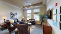 This single-floor recreation center offers a thoughtfully designed communal space with distinct activity zones that cater to a variety of leisure interests. The layout features a generously sized main area anchored by a full-sized pool table and a foosball table, providing ample space for games and social gatherings. Tray ceilings with crown molding lend an elevated architectural character throughout, while ceiling fans and recessed lighting ensure a comfortable, well-lit environment.

The lounge area is furnished with rich leather sofas, armchairs, and wingback chairs arranged around a central coffee table, creating an inviting setting for conversation or relaxation. A wall-mounted flat-screen television serves as a focal point, complemented by decorative wall art panels nearby. A compact arcade gaming station adds an additional entertainment option.

Multiple large windows with horizontal blinds line the walls, allowing abundant natural light to filter into the space and offering views of the surrounding outdoor greenery. High transom windows further amplify the brightness of the room. Neutral warm-toned walls and patterned carpeting throughout contribute to the space's welcoming ambiance. Additional high-top seating near a window provides a casual spot for smaller gatherings or individual use.