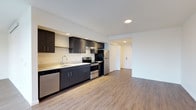 This single-floor space presents a clean, contemporary design with an open-concept layout that seamlessly connects the kitchen and living area. The kitchen is a standout feature, outfitted with dark espresso-toned cabinetry that contrasts elegantly against light countertops and white walls. Stainless steel appliances — including a range with oven, refrigerator, and dishwasher — lend a polished, modern aesthetic, while an under-cabinet range hood adds both function and style. Recessed lighting in the kitchen ceiling provides focused, even illumination throughout the cooking space. Warm, hardwood-style luxury vinyl flooring flows continuously from the kitchen into the open living area, creating a cohesive and inviting atmosphere. The living space is generously proportioned, offering flexible room for a variety of furniture arrangements. A hallway leads to additional areas of the unit, suggesting a thoughtful and functional floor plan. The overall ambiance is modern and understated, with a neutral color palette that provides a versatile backdrop for a range of interior styles. The space feels bright and well-maintained, reflecting quality finishes and attention to detail throughout.