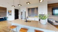 Located at 700 Semmes Ave in Richmond, Virginia, this contemporary amenity lounge offers a thoughtfully designed, open-concept layout across a single floor. Large windows throughout flood the space with natural light and frame sweeping views of the Richmond skyline and lush treeline beyond, creating a bright and inviting atmosphere.

The lounge is divided into distinct yet interconnected zones. A comfortable social seating area features a bold dark accent wall with textured wallcovering, a wall-mounted flat-screen TV, light wood flooring, and a curated mix of neutral and orange upholstered furniture. Adjacent to this is a vibrant game area centered around a billiard table, accompanied by additional seating and direct access to an outdoor balcony, further extending the scenic views.

A welcoming nook near the windows offers a cozy banquette-style orange sofa, a round marble-topped table, and a circular pendant light fixture, ideal for casual conversation or relaxation. Nearby, a sleek reception or co-working station features a wood-paneled accent wall and modern blue seating.

The space also includes a private meeting room enclosed by floor-to-ceiling glass and black-framed partitions. The well-appointed kitchen boasts a large quartz waterfall island with bar seating, white cabinetry, stainless steel appliances, and statement pendant lighting—perfect for community gatherings and events.