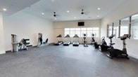 This fitness center offers a clean, functional space designed to support a well-rounded exercise routine. The room features light-toned walls and a durable rubber flooring surface well-suited for high-traffic athletic use. Recessed lighting is distributed evenly across the ceiling, supplemented by two ceiling fans that help maintain a comfortable workout environment. A wide row of windows along the far wall allows natural light to fill the space, creating an open and airy atmosphere.

The cardio section is prominently featured, housing multiple treadmills lined up facing the windows, along with an elliptical trainer and a recumbent bike, offering a variety of low-impact and aerobic exercise options. A strength training machine is positioned along the side wall, providing options for resistance-based workouts. A wall-mounted flat-screen television is installed above the windows, allowing users to stay entertained during their sessions.

The layout is thoughtfully organized to maximize usable floor space while maintaining clear pathways between equipment. The overall design is minimalist and practical, prioritizing functionality and ease of movement. This fitness center provides a well-lit, well-ventilated environment equipped with the essential tools for both cardiovascular and strength training workouts.