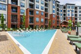 This outdoor amenity space in Marietta, Georgia, is set within a multi-story brick and mixed-material residential community, offering a thoughtfully designed courtyard environment centered around a large, resort-style swimming pool. The pool deck is finished with patterned cobblestone pavers and lined with rows of cushioned green chaise lounges, providing ample space for sunbathing and relaxation. Mature ornamental trees, evergreen shrubs, and colorful plantings frame the perimeter, enhancing the sense of a well-maintained, landscaped retreat.

A prominent timber-framed pavilion anchors one section of the outdoor space, featuring rustic wood beams supported by stacked stone columns. Beneath it, outdoor dining tables with bold red market umbrellas and bar-height seating with grilling stations create a dedicated space for al fresco entertaining. The pavilion overlooks a sports court visible beyond the black iron perimeter fencing, with lush tree lines providing a natural backdrop.

The overall design balances urban architecture with natural greenery, offering a versatile outdoor experience. Cobblestone pathways connect the various zones, unifying the space while maintaining a refined, community-oriented aesthetic throughout.