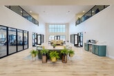 Located at 4005 State Farm Pkwy in Columbia, Missouri, this impressive single-floor amenity facility spans a generous footprint designed to accommodate a wide range of activities and social functions. The centerpiece of the space is a dramatic, double-height entrance lobby bathed in natural light from oversized gridded windows. Light hardwood flooring flows throughout, complemented by crisp white walls accented with bold forest green panels that lend a modern, polished aesthetic. A central lounge area features curated seating arrangements with lush potted ferns in gold-toned planters, creating an inviting gathering point. Adjacent to the lobby, floor-to-ceiling glass partitions reveal a well-equipped fitness center outfitted with cardio machines, strength equipment, and suspension training gear, visible from multiple vantage points throughout the main hall. A dedicated golf simulator room provides a projector-based screen setup with turf hitting mats and built-in cabinetry. The kitchen is appointed with sage green shaker cabinetry, a striking marble-veined stone backsplash and countertops, and modern black fixtures. Additional areas include a glass-enclosed conference or dining room, open lounge seating zones, two hallways, two bathrooms, and a welcoming entrance. An upper-level mezzanine overlooks the main floor, adding architectural depth to the overall design.
