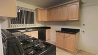 This single-story space includes a well-appointed kitchen that serves as a central feature. The kitchen is equipped with light wood cabinetry featuring arched door detailing, offering generous upper and lower storage throughout. Dark granite-style countertops provide a striking contrast against the warm-toned cabinetry, creating a visually balanced aesthetic. A black gas range with multiple burners anchors the cooking area, complemented by a built-in black dishwasher for added convenience. The sink is positioned beneath a window dressed with vertical blinds, allowing natural light to filter into the space. An additional freestanding cabinetry unit with matching countertop provides extra prep or storage space along the opposite wall. The flooring consists of neutral-toned tile, contributing to the kitchen's clean and practical character. The layout offers a functional workflow with adequate counter space distributed across multiple zones, making it well-suited for everyday cooking and meal preparation. Overall, the kitchen presents a cohesive design with a combination of warm wood tones and dark surfaces.