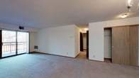 This single-floor residence offers a practical and comfortable layout designed for everyday living. The open living area is generously sized, featuring soft wall-to-wall carpeting, neutral white walls, and a sliding glass door that opens to a balcony, allowing natural light to fill the space. A wall-mounted thermostat and in-unit ventilation grilles are conveniently integrated throughout.

The kitchen is well-appointed with warm honey-toned wood cabinetry extending to the ceiling, offering abundant storage. A full suite of appliances — including a range, dishwasher, and refrigerator — is included, alongside a double basin sink and light countertops. A breakfast bar peninsula connects the kitchen to the living and dining area, which is accented by a classic chandelier overhead.

The bedroom features plush carpeting, a closet with built-in shelving, and a window with blinds that provides views to the outdoors. An additional open closet space is also visible in the layout, adding to the storage options.

The bathroom is clean and functional, featuring a wide vanity with light countertops, ample cabinet storage below, a large mirror with overhead lighting, tile flooring, and a toilet and tub/shower combination. The overall design maintains a consistent, understated aesthetic with warm wood accents and light neutral tones throughout.