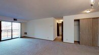This single-floor residence offers a practical and comfortable layout designed for everyday living. The open living area is generously sized, featuring soft wall-to-wall carpeting, neutral white walls, and a sliding glass door that opens to a balcony, allowing natural light to fill the space. A wall-mounted thermostat and in-unit ventilation grilles are conveniently integrated throughout.

The kitchen is well-appointed with warm honey-toned wood cabinetry extending to the ceiling, offering abundant storage. A full suite of appliances — including a range, dishwasher, and refrigerator — is included, alongside a double basin sink and light countertops. A breakfast bar peninsula connects the kitchen to the living and dining area, which is accented by a classic chandelier overhead.

The bedroom features plush carpeting, a closet with built-in shelving, and a window with blinds that provides views to the outdoors. An additional open closet space is also visible in the layout, adding to the storage options.

The bathroom is clean and functional, featuring a wide vanity with light countertops, ample cabinet storage below, a large mirror with overhead lighting, tile flooring, and a toilet and tub/shower combination. The overall design maintains a consistent, understated aesthetic with warm wood accents and light neutral tones throughout.