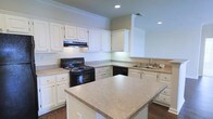 This single-floor residence showcases a thoughtfully designed kitchen that serves as the heart of the home. The kitchen is outfitted with an abundance of white raised-panel cabinetry, providing generous storage and a clean, timeless aesthetic. Light-toned countertops run throughout, offering ample workspace for cooking and entertaining. A central island adds additional prep space and anchors the layout, making the kitchen both highly functional and visually cohesive. Black appliances, including a freestanding gas range with a vent hood above and a dishwasher, create a striking contrast against the white cabinetry. A double-basin sink is positioned along the perimeter cabinetry, accompanied by a classic faucet and tile backsplash detail. Recessed lighting in the ceiling ensures the space is well-illuminated, complemented by crown molding that lends an elevated, finished look to the room. Rich dark hardwood flooring grounds the space and adds warmth. The open sightlines and well-organized layout make this kitchen an efficient and welcoming area, reflecting a practical yet polished design throughout the home.
