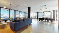 This single-floor space showcases a thoughtfully designed, contemporary interior that seamlessly blends open-plan living with striking architectural details. Expansive floor-to-ceiling glass walls wrap around the main lounge and dining areas, flooding the space with natural light and offering panoramic views of the surrounding urban skyline. Polished concrete flooring extends throughout, lending an industrial yet refined aesthetic to the open layout.

The main lounge area is anchored by a generous sectional sofa and is organized around distinct seating zones, including clusters of accent chairs positioned directly against the glass perimeter to maximize the outdoor views. A dedicated dining area features a sizable table with modern seating beneath a sculptural pendant light fixture, accented by hanging greenery.

The kitchen is a standout feature, designed with bold navy-blue cabinetry and dual islands topped with speckled terrazzo countertops. A vibrant terracotta-toned accent wall with a geometric pattern adds a pop of color and artistic character to the space, complemented by framed artwork. The same geometric wallcovering continues into the entry corridor, where warm-toned concrete flooring and wood-paneled walls create a welcoming transition into the space. A separate lounge room offers additional seating with city views through full-height shaded glazing.