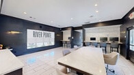 Located at 915 Lovvorn Rd in Carrollton, Georgia, this River Pointe community clubhouse is a well-appointed, single-floor amenity space designed with a clean, modern aesthetic. The interior is defined by large-format marble-look tile flooring, bold dark accent walls, and recessed lighting that creates a bright, professional atmosphere throughout.

The kitchen is a standout feature, equipped with dark wood cabinetry accented by gold hardware, white quartz countertops, a stainless steel range, an undermount sink, and a central island that provides additional prep and gathering space. A full-height tile backsplash in a soft marble finish adds a polished, upscale touch. Additional appliances, including a beverage refrigerator and coffee station, make the kitchen well-suited for events and gatherings.

Adjacent to the kitchen, an open workspace offers a built-in countertop workstation with bar-height seating and computer terminals, ideal for residents seeking a productive environment. A large conference-style table with rolling office chairs anchors the central area, providing flexible space for meetings or collaborative work. A prominent "River Pointe" branded display and large windows allow natural light to filter in, enhancing the inviting, community-focused ambiance of the space.