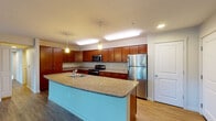 This single-floor residence offers a thoughtfully designed layout that balances functionality and comfort. The centerpiece of the home is the expansive kitchen, which features rich cherry wood cabinetry extending to the ceiling, granite-style countertops, and a suite of stainless steel appliances including a refrigerator, range, and microwave. A large curved kitchen island with a built-in sink and a distinctive teal base provides additional workspace and serves as a natural gathering point. Pendant lighting and recessed ceiling fixtures illuminate the kitchen, enhancing its warm and inviting character. Wood-look flooring runs through the kitchen and flows into the open living area, which is bathed in natural light from multiple windows and a glass-panel door that offers views of the surrounding greenery. The bedrooms are finished with plush carpeting and neutral tones, each featuring windows with exterior views and closet storage — one notably equipped with a mirrored sliding closet door. The bathroom is clean and practical, offering a granite-top vanity with wood cabinetry, a combination tub and shower, and wood-look flooring. A dedicated laundry area is also present within the unit, adding to the overall convenience of this well-rounded residential space.