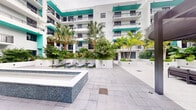 Located at 2001 Hollywood Boulevard in Hollywood, FL, this outdoor courtyard space offers a vibrant and resort-style atmosphere characteristic of South Florida living. The area is defined by its clean, modern aesthetic, anchored by a tiled spa with blue mosaic detailing that serves as a central focal point. Wide, light-toned stone pavers extend across the courtyard, providing a polished and open surface underfoot. Lounge chairs are positioned nearby, creating a relaxed setting ideal for leisure and sunbathing. A sleek pergola structure adds architectural interest and partial shade to one side of the space. Lush tropical landscaping, including tall palm trees, broad-leafed plants, and colorful foliage, is artfully arranged in raised planters throughout, lending a vibrant, garden-like quality to the area. The courtyard is framed by a multi-story residential building featuring crisp white facades, private balconies with dark railings, and bold teal accent banding — a signature design element that gives the property a distinctly contemporary and coastal character. The open-air setting allows for abundant natural light, contributing to the bright and welcoming ambiance of this well-appointed outdoor retreat.