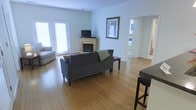 This single-floor home offers a welcoming and open layout designed for comfortable everyday living. The main living area is anchored by warm hardwood flooring that extends throughout the space, creating a cohesive and inviting atmosphere. A classic fireplace with a light-toned mantel serves as a natural focal point, complemented by a television setup and a charming framed piece of wall art that adds a decorative touch.

Natural light pours into the living room through a set of full-length French doors, brightening the crisp white walls and contributing to an airy, open feel. A gray upholstered sofa with nailhead trim is centrally positioned around a wooden coffee table, providing a functional and stylish seating arrangement.

The open-concept design allows for a smooth transition between the living area and adjacent spaces. A bar-height countertop with seating is visible along one side, suggesting a kitchen or dining bar nearby, while a wide doorway leads to additional rooms, hinting at generous overall square footage. The clean, neutral color palette throughout the space creates a versatile backdrop suited to a variety of interior styles. Overall, this home presents a bright, functional, and well-connected layout ideal for both relaxing and entertaining.