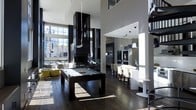 This sophisticated single-floor space showcases a bold contemporary design defined by dramatic double-height ceilings and an open-concept layout that seamlessly connects its key areas. The main living and entertainment zone commands immediate attention with a sleek billiards table positioned beneath a statement pendant light fixture, anchored by rich dark hardwood flooring that runs throughout. A striking black architectural column serves as a dramatic focal point, adding both visual contrast and structural character to the space.

Floor-to-ceiling windows flood the interior with abundant natural light while framing expansive urban views of the surrounding cityscape. The lounge area features modern accent seating in vibrant yellow-green tones, providing a lively contrast against the predominantly monochromatic palette of black, white, and grey.

The open kitchen is fully integrated into the overall layout, outfitted with high-end appliances, ample cabinetry, and a bar-height counter with seating for casual dining. Pendant lighting above the counter zone adds warmth and definition to this functional area. A spiral staircase is partially visible, adding an elegant architectural detail. The overall ambiance is polished and urban, reflecting a refined modern aesthetic throughout.