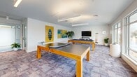 Located at 5832 Coastal Pointe Dr in Virginia Beach, VA, this single-floor community amenity space offers a well-appointed and thoughtfully designed environment for socializing, recreation, and productivity. The layout is divided into distinct functional zones that flow seamlessly into one another. A bright dining and gathering area anchors one end of the space, featuring large-format tile flooring, floor-to-ceiling glass walls, and sliding doors that open directly onto an expansive outdoor terrace with patio seating and sweeping views of the surrounding community. Adjacent to this is a casual lounge area furnished with plush seating, warm-toned carpet tiles, curated artwork, and a wall-mounted TV — ideal for relaxed gatherings or quiet downtime. A dedicated game room provides recreational amenities including ping-pong and billiards tables beneath sleek linear pendant lighting, accented by bold wall art. A co-working or collaborative area is equipped with a large communal table, modern chairs, warm wood-paneled walls, and direct elevator access. Glass-partitioned rooms offer semi-private spaces for focused work or small meetings. Throughout, the design is characterized by clean white walls, abundant natural light, and contemporary furnishings, creating a bright, welcoming, and versatile community hub.