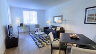 This single-floor space offers a well-designed open-plan layout that seamlessly integrates the living and dining areas into one cohesive environment. The interior is finished with smooth, light-toned hardwood-style flooring that extends throughout, contributing to a clean and modern aesthetic.

The living area is anchored by a dark sofa accented with decorative pillows, complemented by a geometric-patterned area rug in shades of grey, yellow, and white. A round white coffee table sits at the center, while a framed wall piece and a floor lamp add visual interest. A window along one wall allows natural light to fill the space, with views of greenery outside enhancing the bright, airy atmosphere.

The dining area features a dark rectangular table paired with upholstered white chairs, creating a refined contrast that ties in with the overall color palette. A small workspace or accent table near the window adds functional versatility to the layout.

The walls are painted a crisp, neutral white, amplifying the sense of openness. Overall, the space presents a contemporary and well-appointed interior suited for comfortable everyday living.