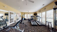This fitness center offers a comprehensive workout environment designed to support a wide range of exercise routines. The space is spread across a single floor and is finished with durable patterned carpet flooring throughout. Warm yellow walls and an abundance of large, blinds-fitted windows allow generous natural light to fill the room, creating an energizing atmosphere. Ceiling fans are positioned throughout to maintain airflow and comfort during workouts.

The cardio section is well-appointed with multiple treadmills, an elliptical trainer, and a stationary bike, complemented by a wall-mounted flat-screen television for entertainment during exercise. A wall clock keeps users on track with their sessions.

The strength and functional training area features a multi-station cable machine, a weight bench with free weights, a dumbbell rack, and a stability ball, catering to a full-body training regimen. TRX-style suspension straps and resistance tools are also available. Large mirrors line one wall, enhancing the sense of space and allowing users to monitor their form.

Workout reference charts are posted near the entrance, and French-style glass-panel doors connect sections of the space. Motivational wall decals add an inspiring touch to the overall environment. The fitness center is well-lit with recessed lighting supplementing the abundant natural light from the surrounding windows.