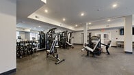 Located at 857A Federal Rd in Brookfield, Connecticut, this fitness center offers a well-appointed, single-floor workout facility designed to support a full range of exercise routines. The space is thoughtfully organized to accommodate both strength training and cardiovascular equipment, providing ample room to move freely between stations.

The strength training area is stocked with a variety of cable machines, multi-functional weight stations, and benches, complemented by a dedicated free weights section with dumbbells and barbells. Full-length mirrors line one wall, enhancing the sense of openness while allowing users to monitor their form. Structural columns are integrated into the layout without disrupting the flow of the space.

The cardio zone features treadmills, ellipticals, and stationary bikes, positioned along a bank of large windows that frame views of the surrounding natural landscape. This abundance of natural light creates an energizing and inviting atmosphere throughout the facility.

Recessed lighting throughout the ceiling ensures bright, even illumination across all areas. The durable, rubber-speckled flooring provides both safety and comfort underfoot. Additional amenities include open storage shelving and a wall-mounted television. The clean, neutral color palette of white and gray walls contributes to the facility's modern, professional aesthetic.
