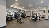 Located at 857A Federal Rd in Brookfield, Connecticut, this fitness center offers a well-appointed, single-floor workout facility designed to support a full range of exercise routines. The space is thoughtfully organized to accommodate both strength training and cardiovascular equipment, providing ample room to move freely between stations.

The strength training area is stocked with a variety of cable machines, multi-functional weight stations, and benches, complemented by a dedicated free weights section with dumbbells and barbells. Full-length mirrors line one wall, enhancing the sense of openness while allowing users to monitor their form. Structural columns are integrated into the layout without disrupting the flow of the space.

The cardio zone features treadmills, ellipticals, and stationary bikes, positioned along a bank of large windows that frame views of the surrounding natural landscape. This abundance of natural light creates an energizing and inviting atmosphere throughout the facility.

Recessed lighting throughout the ceiling ensures bright, even illumination across all areas. The durable, rubber-speckled flooring provides both safety and comfort underfoot. Additional amenities include open storage shelving and a wall-mounted television. The clean, neutral color palette of white and gray walls contributes to the facility's modern, professional aesthetic.