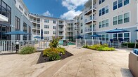 Located at 715 US Highway 46, this outdoor amenity space is centered around a generously sized resort-style swimming pool that serves as the focal point of the courtyard. The pool deck is finished in large-format stone-toned pavers, providing a clean and durable surface that extends across the entire outdoor area, offering ample room for lounging and socializing.

Surrounding the pool is a well-curated selection of outdoor furniture, including modern sling-style chaise lounges and seating arrangements shaded by blue market umbrellas, creating comfortable areas for relaxation throughout the day. The courtyard is enclosed on multiple sides by a multi-story contemporary building, characterized by its light gray and white horizontal siding, private balconies, and large windows that reflect the outdoor environment.

Thoughtful landscaping enhances the space, with planting beds featuring ornamental grasses, black-eyed Susans, and decorative shrubs bordered by fresh mulch. A pergola structure is also present, adding architectural interest and additional shaded gathering space. A white metal perimeter fence frames the pool area cleanly, and subtle exterior lighting fixtures are positioned throughout the courtyard. The overall design prioritizes an open, airy atmosphere with clear sightlines and a cohesive modern aesthetic that blends functionality with resort-inspired comfort.