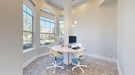 Located at 920 Yorkmont Ridge Lane in Fort Mill, SC, this single-floor space serves as a well-appointed business center designed for productivity and collaboration. The main workspace is defined by soaring ceilings and dramatic floor-to-ceiling windows with arched transoms that flood the room with natural light, offering views of the surrounding community. Elegant pendant lighting clusters hang above a central round white worktable, which is equipped with desktop computers and ergonomic rolling chairs, creating an inviting and functional setting for residents or professionals. The textured, patterned carpet adds warmth and visual interest throughout. Structural columns frame the workspace area, lending an architectural character to the room. The entryway is clean and minimal, featuring large glass-paneled doors that connect the interior to an outdoor brick-paved porch, enhancing the sense of openness. A fire safety panel and climate control are discreetly mounted near the entrance, reflecting thoughtful building management. Crown molding and crisp white baseboards throughout contribute to a polished, professional finish. The overall ambiance is light, airy, and modern, offering a comfortable environment well-suited for focused work or collaborative meetings.