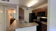 This single-story space centers around a well-appointed kitchen designed for practical everyday use. The kitchen features rich, dark-toned wood cabinetry with shaker-style upper and lower cabinets, providing ample storage throughout. A peninsula extends from the main kitchen layout, topped with a granite-look laminate countertop and equipped with a double-basin stainless steel sink, creating a natural division between the kitchen and surrounding areas. Black appliances, including a refrigerator, electric range, and over-the-range microwave, complement the dark cabinetry for a cohesive aesthetic. Overhead, a large flush-mount fluorescent light fixture provides bright, even illumination across the workspace. The kitchen connects openly to adjacent spaces, with a glimpse into what appears to be a bathroom featuring a vanity with mirror. Neutral-toned walls and a textured ceiling contribute to a clean, understated interior. A ceiling vent indicates a central HVAC system. The overall layout prioritizes functionality and flow, making the kitchen the focal point of this efficiently organized single-floor space.