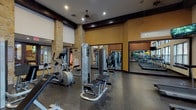 This fitness center offers a comprehensive workout environment designed with both functionality and aesthetics in mind. The space is defined by striking natural stone columns that rise toward elevated ceilings accented with dark wood beams, creating a warm yet energetic atmosphere. Recessed lighting and a ceiling fan ensure the space remains well-lit and comfortable during use.

The floor is finished with durable rubberized dark flooring, suitable for heavy equipment and high-traffic use. Large mirrored panels line one wall, visually expanding the space while allowing users to monitor their form during exercise. On the opposite side, a row of oversized windows fitted with horizontal blinds floods the cardio zone with natural light and provides outdoor views, enhancing the overall experience.

Equipment is thoughtfully arranged throughout the space. The cardio section includes multiple treadmills and elliptical machines positioned along the windowed wall, accompanied by a wall-mounted flat-screen television. The strength-training area features a variety of cable machines, weight benches, and a free weights rack with dumbbells. A vending machine is also available for added convenience, making this fitness center a fully equipped facility for a well-rounded exercise routine.
