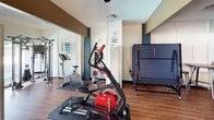 This single-floor amenity space seamlessly blends a fitness area with a recreational lounge, offering a well-rounded environment for both exercise and leisure. The fitness zone is equipped with a variety of cardio and strength-training machines, including an elliptical trainer, stationary bike, and a multi-function cable weight machine, all positioned on protective floor mats over warm hardwood-style flooring. A folded ping-pong table stands ready for use, adding a playful element to the workout area. Large sliding glass doors at one end of the fitness space allow natural light to flood the room and provide views to an outdoor area, creating an open, airy atmosphere. Teal-painted structural columns and rich brown accent beams provide a modern, color-blocked aesthetic throughout the space. The adjoining recreation lounge features a full-size pool table as its centerpiece, complemented by a wall-mounted flat-screen television and multiple high-top bistro tables with bar stools — ideal for casual social gatherings. Textured ceilings with flush-mount and ceiling fan lighting fixtures illuminate both zones evenly. A corridor near the lounge leads to exterior access, making this a conveniently connected communal facility overall.