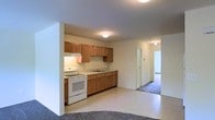 This single-floor space features a practical, open-concept layout that connects the kitchen directly to the surrounding living areas. The kitchen is equipped with warm honey-toned wood cabinetry spanning both upper and lower storage, a white electric range, a single-basin sink with under-cabinet lighting, and light-colored countertops that provide a clean, functional workspace. The flooring in the kitchen area is a smooth, light-toned vinyl or linoleum, offering easy maintenance.

A half-wall partition subtly defines the kitchen from the adjacent living space, which transitions into plush wall-to-wall carpeting that extends throughout the remainder of the unit. Flush-mount ceiling fixtures provide consistent overhead lighting throughout the space. A hallway visible from the main area leads to additional rooms, with a door at the end hinting at a private bedroom or bathroom. The walls throughout are finished in a neutral off-white tone, creating a bright, clean atmosphere. The overall design is straightforward and functional, prioritizing practical storage and efficient use of space across the single-floor layout.