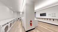Located at 2032 Worthington Cir in Santa Clara, California, this single-story shared laundry facility offers a thoughtfully designed space that balances functionality with comfort. The interior features light wood-look flooring throughout, crisp white walls, and recessed linear LED lighting that creates a consistently bright and clean atmosphere.

The laundry area is well-appointed with a generous row of front-loading washers and dryers along one wall, along with additional top-loading machines and utility sink stations, providing ample capacity for high-volume use. A structural column divides the space, with a fire extinguisher station conveniently mounted nearby. The machines are arranged for easy access and efficient flow.

A dedicated lounge area adjacent to the laundry zone enhances the user experience, featuring a wall-mounted flat-screen television, a long wall-mounted folding counter with bar stools for sorting or working, and comfortable seating including tan leather and navy upholstered chairs. A large window fitted with roller shades allows natural light and views of the surrounding greenery. An accent wall painted in soft sage green, a wall-mounted split HVAC unit, and decorative artwork complete the inviting atmosphere. Double entry doors provide easy access to and from the facility.