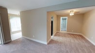 This single-floor residential space presents a clean, move-in-ready interior defined by its neutral warm beige walls and soft wall-to-wall carpeting throughout. The open layout creates a sense of spaciousness, with the main living area flowing naturally into connecting rooms via wide doorways and a hallway. White baseboards provide a crisp contrast against the warm wall tones, contributing to a tidy and well-maintained aesthetic. The ceiling features a ceiling fan with a light fixture, providing both ventilation and ambient illumination. The open passage between rooms allows for an easy, connected flow, making the layout practical and versatile. A hallway visible from the main living area leads to additional rooms, with one doorway revealing a glimpse of a darker-toned room and another leading further down the corridor to a closed door, suggesting multiple private spaces. The overall design is straightforward and functional, with consistent finishes across the visible areas, including uniform carpeting and wall color, lending the space a cohesive and unified appearance. The interior is unfurnished, allowing prospective occupants to envision a variety of layout configurations suited to their needs.