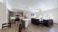 This single-floor unit showcases a thoughtfully designed open-concept layout that seamlessly connects the kitchen, dining, and living areas. The kitchen is a standout feature, equipped with slate-gray shaker-style cabinetry, quartz countertops, stainless steel appliances including a range, microwave, dishwasher, and refrigerator, and a central island with pendant lighting overhead. Wood-look flooring extends throughout the main living areas, adding warmth and visual continuity.

The dining area includes a built-in desk nook with matching gray cabinetry, offering a practical workspace alongside the dining table. The living room benefits from generous natural light streaming through large windows fitted with vertical blinds, a ceiling fan, and an electric fireplace that adds a cozy focal point to the space.

The bedrooms are carpeted and comfortably sized, each featuring ceiling fans. One bedroom includes a walk-in closet, while another accommodates twin beds and receives ample natural light. Two bathrooms are present, both featuring gray vanity cabinetry, quartz countertops, and shower-tub combinations, maintaining the unit's consistent modern aesthetic throughout.