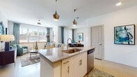 This well-appointed single-floor residence offers a thoughtfully designed open-concept layout that seamlessly connects the kitchen, dining, and living areas. The kitchen is a standout feature, showcasing granite countertops, a central island with an integrated sink, stainless steel appliances including a refrigerator, microwave, and range, white cabinetry with gold-toned hardware, and a herringbone tile backsplash. Stylish pendant lights hang above the island, adding a contemporary touch. The kitchen flows effortlessly into the bright and airy living room, which is filled with natural light from multiple windows dressed with patterned curtains and blinds. A ceiling fan completes the comfortable atmosphere of the living space. The bedroom features a soothing teal accent wall, plush carpeting, and ample natural light, creating a calm and restful retreat. The bathroom is finished with granite vanity counters, white cabinetry, and a full tub-shower combination with tile surround. A dedicated laundry area houses a full-size washer and dryer with built-in shelving above. Throughout the residence, light-toned flooring, neutral walls, and cohesive teal and gold accents create a modern, harmonious aesthetic.