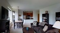 This single-floor residence is designed with an open-concept layout that integrates the living, dining, and kitchen areas into one cohesive, functional space. The kitchen features warm wood cabinetry, stainless steel appliances including a refrigerator and microwave, and a peninsula with a dark countertop that serves as a natural divider between the kitchen and the rest of the living area. Track lighting runs along the ceiling above the kitchen, providing focused illumination over the workspace.

Adjacent to the kitchen, the dining area accommodates a rectangular table with seating for several guests, positioned near a window that allows natural light to filter into the space. The living area is comfortably furnished with a large, dark upholstered sofa accented with decorative pillows, anchored by a round wooden coffee table. A tall bookcase filled with books and decorative items lines one wall, adding a personal and functional touch to the room.

The space features neutral-toned carpeting and light-colored walls, contributing to a bright and welcoming atmosphere. The overall layout is practical and inviting, making efficient use of the available space.