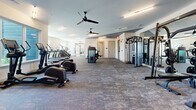 Located at 1100 Rea Dr. in Gainesville, GA, this modern fitness center offers a spacious, single-floor layout designed to accommodate a full range of workout routines. The space is thoughtfully organized into distinct functional zones, providing ample room for both cardio and strength training activities without overcrowding.

The cardio zone features multiple treadmills, stair climbers, and elliptical machines positioned along a windowed wall, allowing natural light to fill the area and offering outdoor views to keep workouts refreshing. Overhead ceiling fans provide additional air circulation throughout.

The strength training area is equally well-appointed, with a full rack of dumbbells, a multi-station cable and power rack system, adjustable benches, and various specialty machines. Large floor-to-ceiling mirrors line the walls in the strength zone, enhancing the sense of openness and allowing users to monitor their form.

The interior design is clean and contemporary, with white walls, recessed and linear LED lighting, and durable, neutral-toned carpet flooring throughout. A wall-mounted television adds an entertainment element to the space. Multiple access doors ensure convenient entry and exit. The overall ambiance is bright, functional, and professionally outfitted, making this fitness center a comprehensive amenity for those seeking a well-rounded exercise facility.