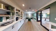 This single-floor amenity space is thoughtfully designed with a cohesive aesthetic that blends sophistication and comfort. Throughout, deep forest green accent walls are paired with crisp white wainscoting and crown molding, creating a refined, club-like atmosphere. Natural light filters in through multiple windows dressed with patterned curtains, enhancing the welcoming ambiance.

The lounge area serves as a central gathering space, featuring plush emerald green velvet armchairs arranged around a round coffee table beneath a classic pendant light fixture. The carpeted flooring adds warmth, while framed artwork and decorative accessories contribute to an inviting atmosphere.

Adjacent to the lounge, the game room commands attention with a rustic pool table as its centerpiece, set atop wood-look flooring beneath an elegant coffered ceiling. High bar-style seating lines the walls, and multiple windows provide ample natural light.

A dedicated business center offers a built-in workstation with dual computer setups, complemented by white cabinetry and open shelving for practical storage. Black-framed glass doors connect the various areas, maintaining an open flow throughout the space while providing visual continuity and a modern architectural accent.