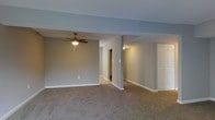 This single-floor space presents a clean, open layout finished in a calm, neutral palette. The main living area is finished with wall-to-wall plush carpeting in a warm taupe tone, providing a comfortable underfoot feel throughout. Walls are painted in a soft gray, lending a modern yet understated aesthetic that pairs well with the white baseboards and trim detailing.

A ceiling fan with a built-in light fixture is centrally mounted, offering both air circulation and ambient lighting to the main living area. Structural columns subtly define zones within the open floor plan without fully closing off the space, maintaining an airy, connected feel.

A hallway extends from the main area, leading toward additional rooms and suggesting a practical, well-organized layout. A white paneled door is visible to one side, hinting at closet or utility storage. Wall-mounted lighting fixtures provide supplemental illumination along the corridor.

Overall, the space is move-in ready with a neutral, versatile interior that serves as an ideal blank canvas for a variety of furnishing styles and personal décor choices.