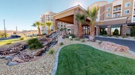 This thoughtfully designed community space makes a strong first impression with its grand exterior entrance, framed by stone-clad columns, a curved metal canopy, and floor-to-ceiling glass doors that flood the interior with natural light. Tall palm trees and manicured desert landscaping surround a decorative rock waterfall feature, lending the approach a resort-like ambiance.

Inside, the single-floor layout opens to a welcoming lounge area finished with warm wood-look flooring and crisp white walls. A stylish accent wall clad in textured gray tile serves as the focal point, anchoring a sleek dark-navy built-in credenza topped with light stone countertops. The credenza hosts a refreshment station, offering residents or visitors a convenient and attractive amenity. A circular framed mirror and paired wall sconces complete this vignette with a polished, modern touch.

The lounge itself is furnished with patterned upholstered seating arranged around a central accent table, creating a comfortable and social gathering spot. Wide corridors extend beyond the lobby, providing clear sightlines throughout the space. The overall design balances contemporary finishes with approachable warmth, making this an ideal hub for community interaction and everyday convenience.