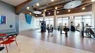 Located at 1201 W Grove St in Boise, Idaho, this modern fitness facility spans a generous single floor and offers a comprehensive, well-appointed environment for health and wellness. The space is defined by its contemporary design, featuring a combination of light tile flooring, warm wood accents, and bold blue wall tones that create an energetic yet welcoming atmosphere.

The main exercise area is divided into distinct training zones. A large turf-floored section is equipped with treadmills, free weights, kettlebells, medicine balls, a functional training rig with TRX and resistance attachments, and a punching bag — all framed by floor-to-ceiling mirrors and expansive windows that flood the space with natural light and outdoor views. An adjacent area with hardwood flooring houses additional strength and cardio machines, including cable systems, rowing machines, and stability equipment, alongside mounted televisions for entertainment.

A standout feature is the indoor climbing wall, offering a vibrant multi-colored bouldering surface beneath soaring ceilings. Nearby, a dedicated open floor space with mattress padding provides room for stretching, yoga, or recovery work.

The facility also includes a stylishly designed lounge and reception area, complete with a wood-paneled planter island filled with lush greenery, built-in seating, a wet bar with beverage refrigeration, and large-scale sports artwork — providing a social and functional gathering point within the facility.