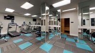 Located at 225 W Third Street in Long Beach, California, this fitness center offers a thoughtfully designed, single-floor workout facility built to support a wide range of exercise routines. The space features a distinctive flooring layout combining grey modular carpet tiles with vibrant teal accent mats, creating clearly defined workout zones throughout the open floor plan.

The main workout area is outfitted with a full rack of dumbbells and medicine balls for strength and functional training, alongside a multi-functional cable machine and adjustable weight bench, providing versatile resistance training options. Large floor-to-ceiling mirrors line the walls, enhancing the sense of space and allowing users to monitor their form during exercise.

A dedicated cardio zone houses treadmills, an elliptical, and a stationary bike, all positioned beneath a wall-mounted television for an engaging workout experience. Red plyometric boxes add a dynamic element for high-intensity interval training.

The facility is well-lit throughout by recessed panel lighting set into a drop ceiling, maintaining a bright and energetic atmosphere. A sauna room with warm wood-paneled walls offers a relaxing recovery option. A small reception or staff area with interior windows provides an oversight view into the main floor. The overall design is clean, modern, and functional, catering to a comprehensive fitness experience.