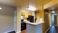 This single-floor space centers around a well-organized kitchen that serves as the heart of the layout. The kitchen is outfitted with a full suite of black appliances, including a refrigerator and an electric range with overhead ventilation, providing a practical cooking environment. Light wood cabinetry lines the upper and lower walls, offering ample storage, while the white countertops provide a clean, contrasting work surface throughout.

A peninsula-style breakfast bar extends into the adjacent living area, creating a natural division between the kitchen and the surrounding space while also offering additional seating and serving options. This open-concept design promotes a sense of connectivity between rooms.

A textured ceiling with flush-mounted overhead lighting ensures the kitchen area is well-lit. Neutral wall tones in warm beige complement the wood cabinetry and contribute to an inviting atmosphere. A doorway visible from the kitchen suggests additional rooms beyond, further extending the functional layout of the space. Built-in shelving near the kitchen provides additional storage and display options, adding to the overall practicality of the unit.