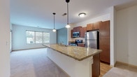 Located at 1200 N 62nd St in Wauwatosa, Wisconsin, this single-floor residence offers a well-designed open-concept layout that seamlessly connects the kitchen and living areas. The kitchen serves as a central highlight, featuring a large granite-topped island with a built-in sink and pendant lighting overhead, rich medium-toned wood cabinetry, and a full suite of stainless steel appliances including a refrigerator, range, and microwave. Hardwood flooring anchors the kitchen space, while plush carpeting extends throughout the living and bedroom areas for added comfort. The open living area is bathed in natural light from generously sized windows that frame views of the surrounding neighborhood, and a set of glass-paned doors provides direct access to an outdoor space. The bedrooms are well-proportioned, each offering soft carpeted flooring, large windows, and closet storage. The bathroom is cleanly appointed with tile flooring, a wood-toned vanity with ample cabinet storage, a large mirror, and a full tub-shower combination. The overall design emphasizes a neutral, contemporary palette, with consistent finishes that create a cohesive and functional living environment throughout the home.