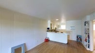 This single-floor space is designed with an open-concept layout that seamlessly connects the living area to the kitchen. The interior is finished in a clean, all-white palette, with paneled walls adding subtle texture to the living space. Light wood laminate flooring runs throughout, contributing warmth and continuity across the open floor plan.

The kitchen is positioned at the far end of the space and features a white peninsula counter that extends outward, providing additional workspace and serving as a natural divider between the kitchen and the living area. White cabinetry lines the upper walls of the kitchen, offering ample storage, while a refrigerator and basic appliances are visible, indicating a fully functional setup. Recessed lighting illuminates the kitchen area, complementing the natural light filtering in through a window.

The living area is spacious and open, with smooth white walls and a clean ceiling that enhance the sense of airiness throughout. The minimalist design approach keeps the space feeling uncluttered and versatile, making it well-suited for a variety of uses. Overall, the space presents a bright, functional, and understated aesthetic.